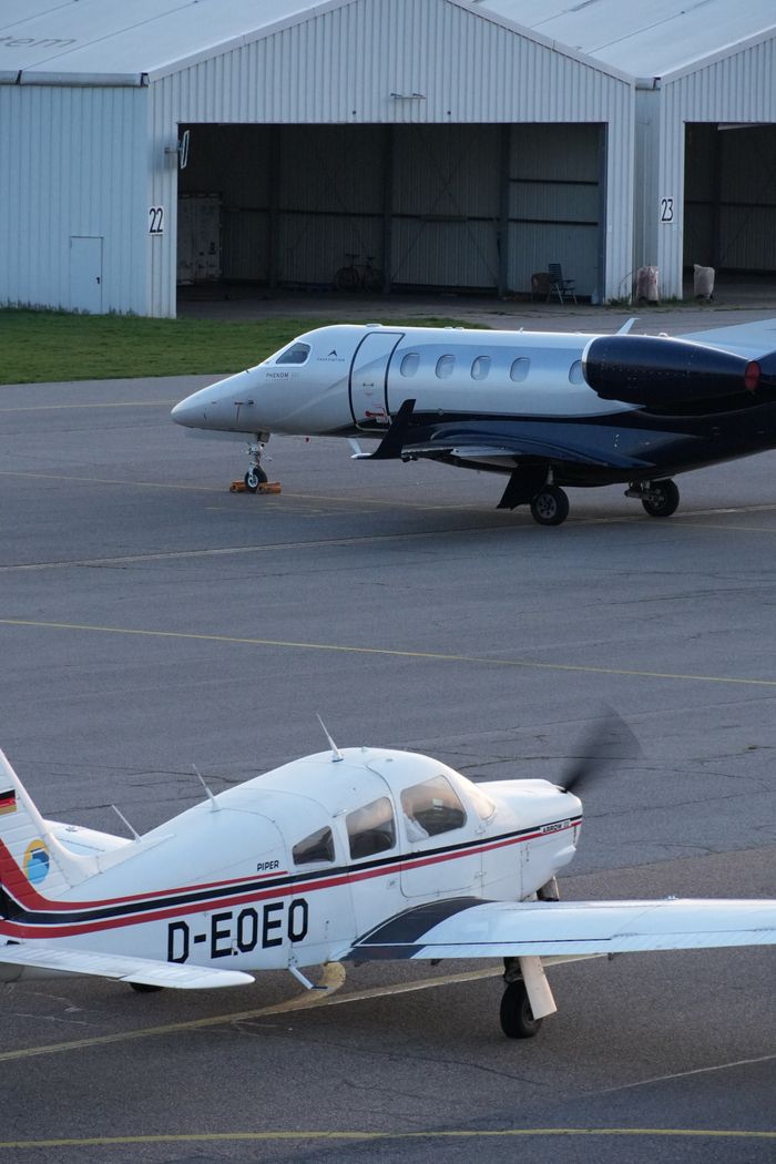About Two private aircraft stationed at a European airfield with hangars in the background.